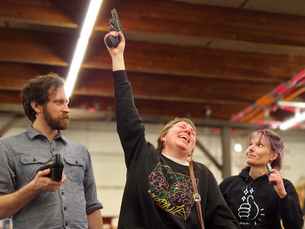 Three adults in a workshop cheer as the center person raises a black tool/prop overhead, smiling while the others look on (left holds a shoe, right holds a small object).