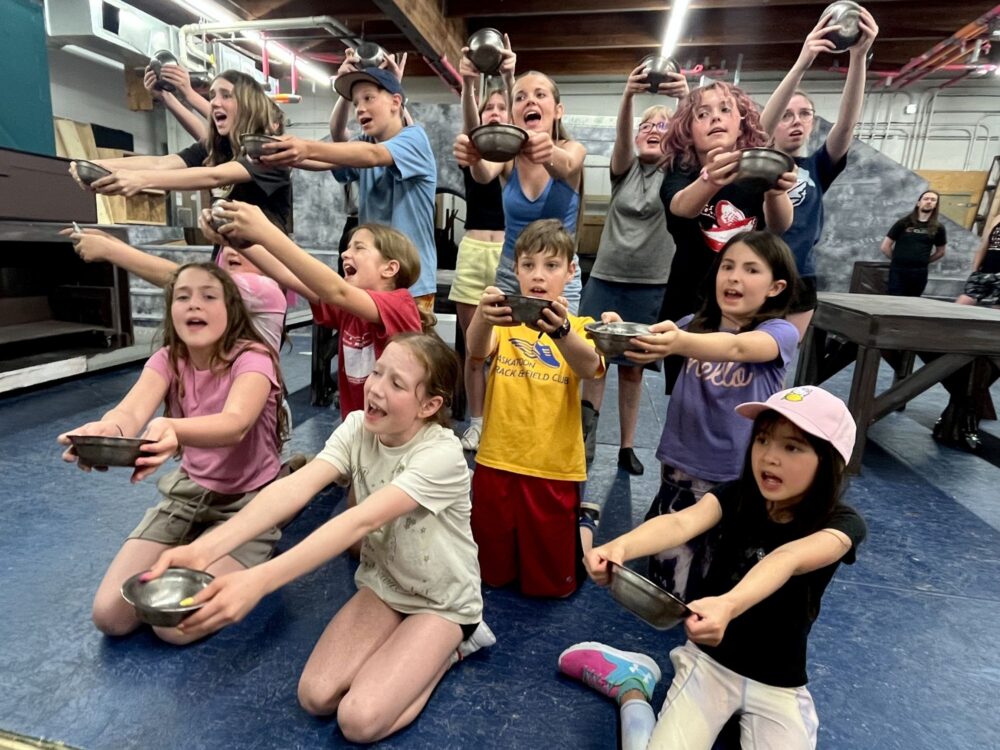 Group of kids kneeling on a blue gym floor, holding metal bowls toward the camera in a playful game inside a gym.