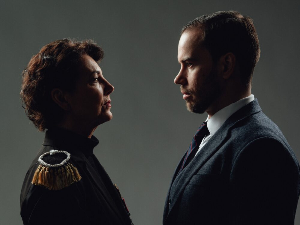 Two adults face each other in profile, a woman in ceremonial uniform with gold epaulettes on the left and a man in a suit and tie on the right.